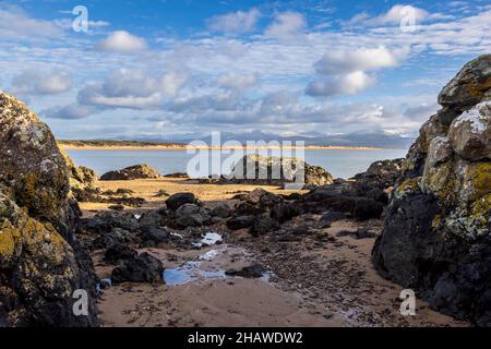 Ein felsiger Strand bei Ebbe auf Ynys Llanddwyn mit den Snowdonia-Bergen über der Bucht, Isle of Anglesey, Nordwales Stockfoto