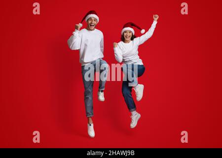 Urlaubsstimmung. Überglücklich Paar Trägt Santa Hats Springen Auf Red Studio Hintergrund Stockfoto