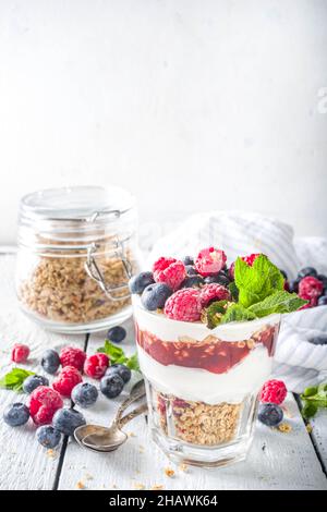 Joghurt parfafait mit Müsli und Beeren. Süßes und gesundes Frühstücksdessert im Glas mit Müsli, Joghurt, Heidelbeeren und Himbeeren. Weißes Holz Stockfoto