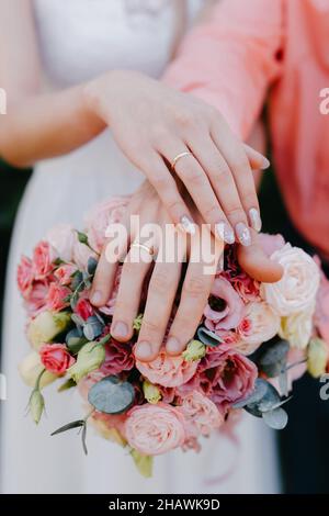HONIGMONDE HÄNDE AUF EINEM HOCHZEITSSTRAUSS AUS BLUMEN. Hände und Ringe auf dem Brautstrauß. Braut und Bräutigam halten sich bei einer Hochzeit mit goldenen Eheringen an die Hand Stockfoto