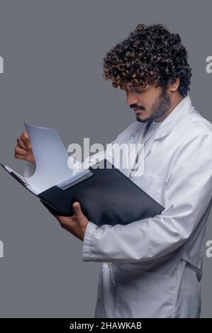 Konzentrierter indischer Arzt mit lockigen Haaren prüft Dokumente Stockfoto