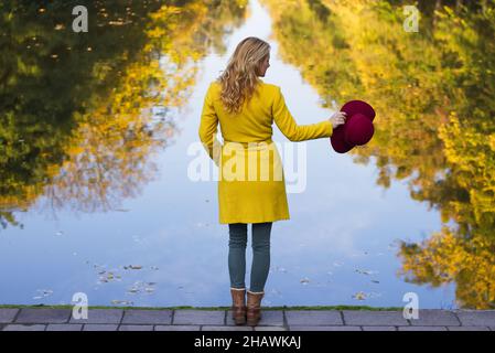 Modische Frau, die im Park über dem See steht und einen roten Hut hält. Spiegelung der Herbstbäume im Wasser. Trendige Frau trägt Wollmantel. Stockfoto