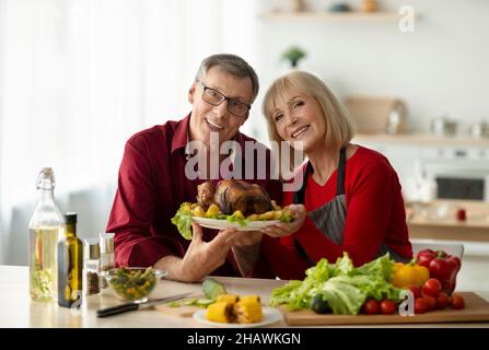 Porträt eines glücklichen älteren Paares, das Gericht mit schmackhaftem gebratenem truthahn für die Weihnachtsfeier der Familie in der Küche hält Stockfoto