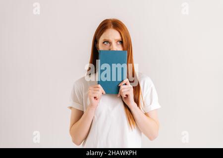 Das Studioporträt einer frustrierten jungen Studentin verbirgt das Gesicht hinter dem Notebook und blickt auf die Kamera, die auf einem weißen, isolierten Hintergrund steht. Stockfoto