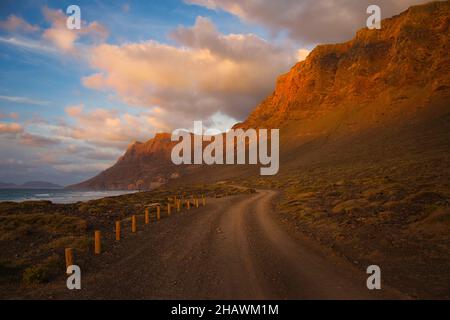 Sonnenuntergang in der Nähe von Famara Beach, Lanzarote, Spanien. Ein Pfad entlang des Meeres und der Klippen. Einige Wolken in einem blauen Himmel. Das letzte Sonnenlicht scheint auf die absichtserklärung Stockfoto