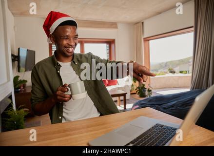 Junger erwachsener schwarzer Mann mit Weihnachtshut, lacht über Witz, während er online telefonieren muss. Sitzen an der Küchentheke mit Laptop. Stockfoto