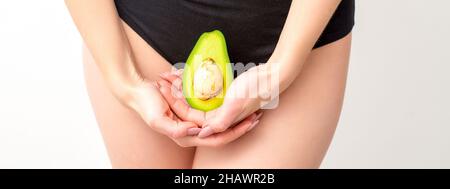 Eine junge Frau hält die Hälfte einer frischen Avocadofrucht vor weißem Hintergrund dicht am Bauch, gesunde Ernährung und Schwangerschaftskonzept Stockfoto