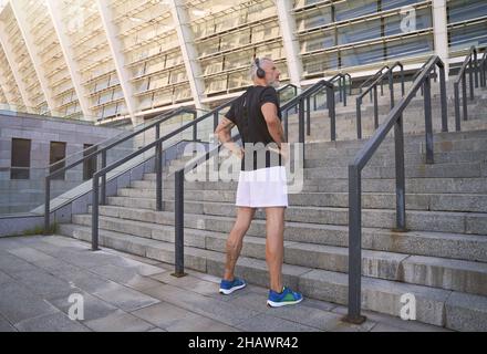 Eine Ganzkörperaufnahme des Sportlers in Kopfhörern, der wegschaut, während er im Freien steht und sich für das morgendliche Workout in der Stadt vorbereitet Stockfoto
