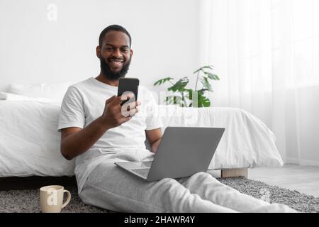Fröhlich lächelnder, hübscher junger afroamerikanischer Mann mit Bart, der am Laptop und Telefon arbeitete und im Schlafzimmer chattet Stockfoto