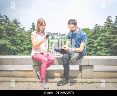 Zwei junge Menschen mit Masken sitzen auf den Zementblöcken in einem Park und schauen auf ihre Telefone. Soziale Medien und Sozialisierung während Pandemien. Stockfoto