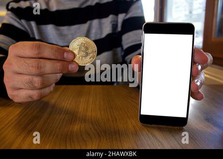 Mann, der eine goldene Bitcoin-Münze hält und ein Mobiltelefon mit leerem weißen Bildschirm zeigt, während er in einem modernen Café sitzt. Stockfoto