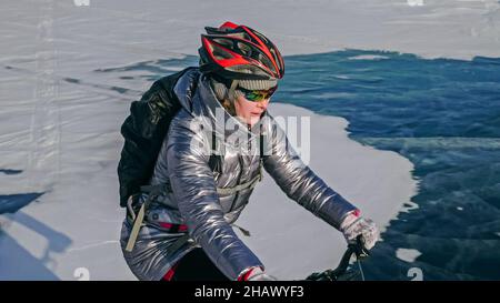 Die Frau fährt Fahrrad auf dem Eis. Mädchen ist in einer silbrigen Daunenjacke, Radrucksack und Helm gekleidet. Eis des gefrorenen Baikalsees. Reifen auf bik Stockfoto