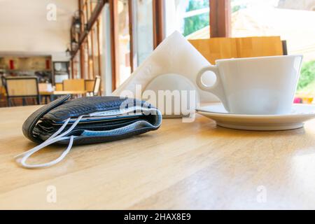 Schwarze Geldbörse mit Gesichtsmaske innen mit weißer Tasse, Servietten in einem Halter um im Café ohne Menschen. Sicherheitsregeln und -Bestimmungen in der Post-Corona p Stockfoto