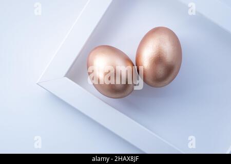 Paar goldene Ostereier mit geometrischem Rahmen auf weißem Hintergrund. Minimalistische Komposition zu Ostern. Flach liegend, Draufsicht, Kopierbereich Stockfoto