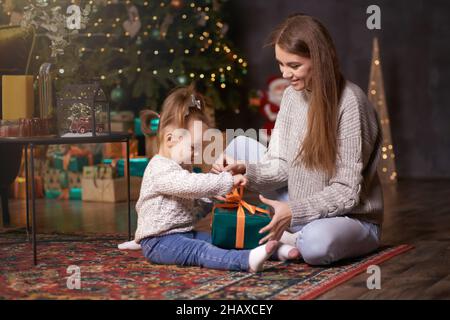 Glückliche Mutter mit Tochter in der Nähe Weihnachtsbaum packt Geschenk. Gemeinsam Spaß haben. Junge Familie im festlichen Zuhause. Ehrlicher, wahrer Moment. Gut Stockfoto