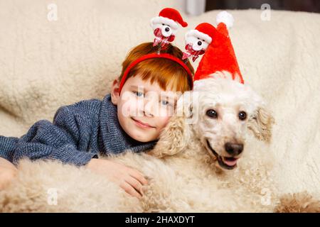 Kind und Hund für Neujahr und weihnachtskonzept. Ein kleiner Junge umarmt einen großen hellen königlichen Pudelhund. Stockfoto