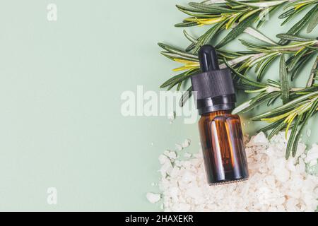 Rosmarin ätherisches Öl Flasche neben frischen Zweigen von Rosmarin und Meersalz in großen Kristallen auf grünem Hintergrund mit Kopierraum, flach gelegt Stockfoto