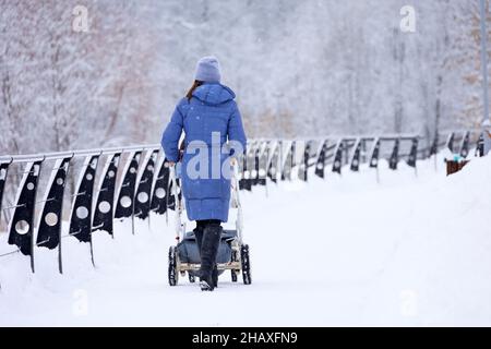 Mädchen in blauem Mantel und Stiefeln, die mit einem Kinderwagen im Winterpark spazieren gehen. Schneewetter, Konzept der Mutterschaft, alleinerziehende Mutter mit Kinderwagen Stockfoto