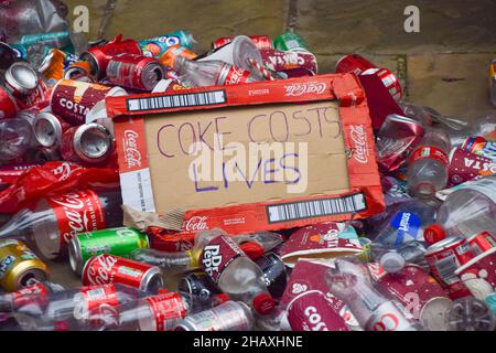 London, Großbritannien. 15th Dez 2021. Während der Demonstration wird das Plakat "Koks kostet Leben" aus einer Kokebox zwischen Plastikflaschen und Blechdosen gesehen.Aktivisten des Extinction Rebellion warfen Plastikflaschen und Blechdosen vor dem Coca-Cola European Partners Hauptquartier in Uxbridge ab. Neben der globalen Umweltverschmutzung beschuldigen die Aktivisten den Limonengiganten der Manipulation, Menschenrechtsverletzungen und Gier der Unternehmen. Kredit: SOPA Images Limited/Alamy Live Nachrichten Stockfoto