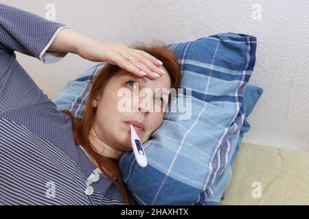 Eine kranke Frau liegt im Bett und misst ihre Temperatur, eine virale Erkältung und Kopfschmerzen. Stockfoto