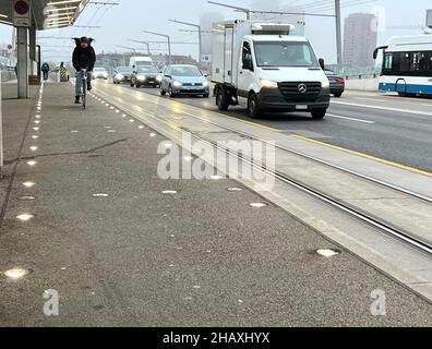 Mann, der in Zürich in der Schweiz auf einer mit Lichtern markierten Fahrradspur auf dem Boden Fahrrad fährt. Parallel gibt es Schienen für die Straßenbahn und befahrene Straße für Autos. Stockfoto