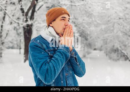 Porträt eines jungen Mannes im verschneiten Winterpark, der eine Jeansjacke trägt und sich beim Spazierengehen im Freien kalt fühlt. Kerl wärmende Hände mit Atem Stockfoto