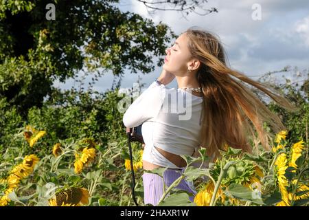 Schönes Teenager-Mädchen, das auf einem Sonnenblumenfeld steht und mit ihren Haaren spielt, Frankreich Stockfoto