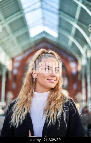 Niedriger Winkel der jungen Frau mit blondem lockiges Haar, das schwarzen Mantel trägt, steht in einem modernen Gebäude Stockfoto