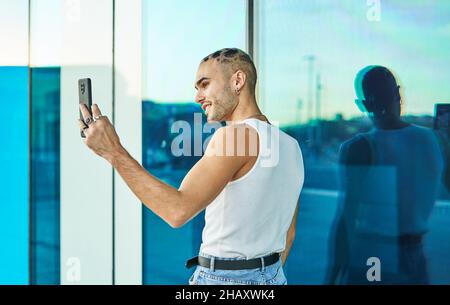 Rückansicht des positiven jungen Mannes mit stilvollem exzentrischem Aussehen, der Selbstporträt auf dem Handy in der Nähe einer spiegelnden blauen Glaswand aufnahm Stockfoto