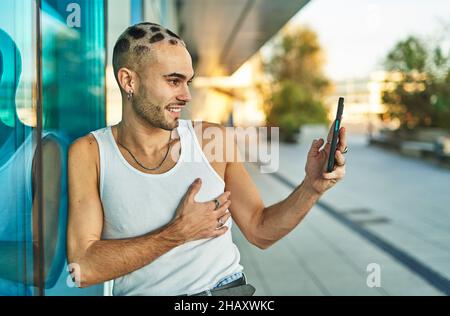 Positiver junger Mann mit stilvollem exzentrischem Aussehen, der Selbstporträt auf dem Handy in der Nähe einer spiegelnden blauen Glaswand aufnahm Stockfoto