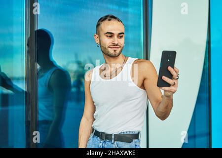 Positiver junger Mann mit stilvollem exzentrischem Aussehen, der Selbstporträt auf dem Handy in der Nähe einer spiegelnden blauen Glaswand aufnahm Stockfoto