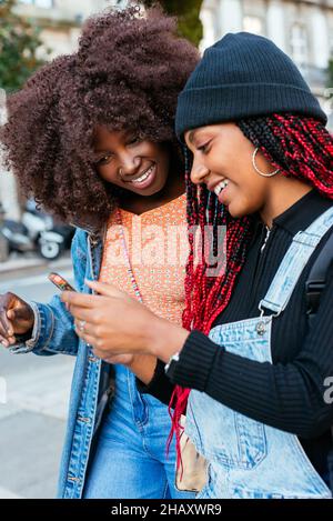 Junge ethnische Freundinnen mit Afro-Frisuren konzentrieren sich, während sie gemeinsam auf einer verschwommenen Straße in der Stadt stehen und Mobiltelefone durchsuchen Stockfoto