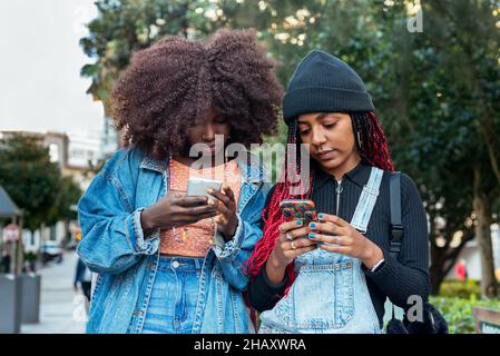 Junge ethnische Freundinnen mit Afro-Frisuren konzentrieren sich, während sie gemeinsam auf einer verschwommenen Straße in der Stadt stehen und Mobiltelefone durchsuchen Stockfoto