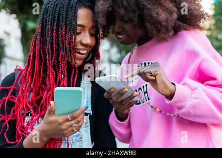 Junge ethnische Freundinnen mit Afro-Frisuren konzentrieren sich, während sie gemeinsam auf einer verschwommenen Straße in der Stadt stehen und Mobiltelefone durchsuchen Stockfoto