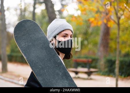 Junge Frau in schwarzer Jacke mit Skateboard am Hals, die auf dem Park steht und eine Gesichtsschutzmaske trägt Stockfoto