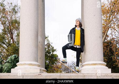 Niedriger Winkel des Ganzkörpers junge positive Frau in lässiger Kleidung, die auf dem Skateboard zwischen Steinsäulen im Park steht Stockfoto