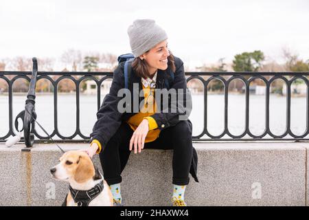 Lächelnde junge Hündin in legerem Outfit in gestricktem Hut und mit Backpacker, die auf der Barriere des Flussufers sitzt und einen liebkosen reinrassigen Hund streichelt Stockfoto