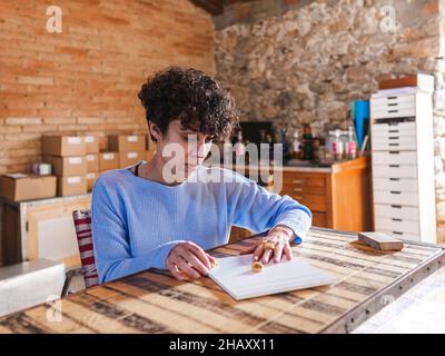 Hispanische Juwelierin sitzt am Tisch und versucht an Ringen, die in der Licht-Werkstatt auf weißer Plakette angebracht sind Stockfoto
