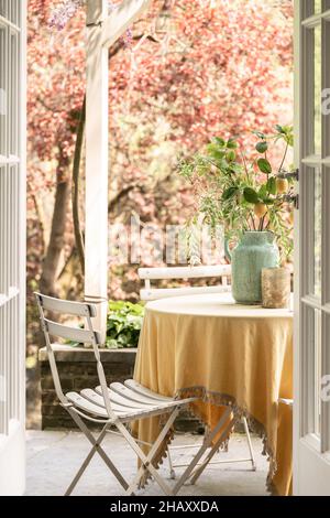 Interieur von gemütlichen Balkon mit runden Tisch mit Tischdecke und Keramikvase mit grünen Pflanzen und Stühlen in der Nähe blühenden Kirschbaum bedeckt Stockfoto
