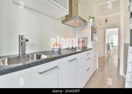 Interieur der modernen hellen schmalen Küche mit weißen Schränken mit eingebautem Backofen und Zähler mit Waschbecken und Herd auf Parkettboden platziert ausgestattet Stockfoto