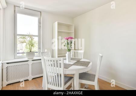Die Inneneinrichtung der kleinen weißen Essecke ist mit einem runden Holztisch mit Stühlen ausgestattet, der auf Parkettboden in der Nähe des weißen Schranks und Fensters platziert und dekoriert ist Stockfoto