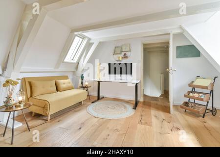 Stilvolles Interieur des Wohnzimmers im Dachgeschoss mit komfortablem gelben Sofa und einem hellen Tisch mit Monitor mit schwarzem Bildschirm in einer modernen Wohnung Stockfoto