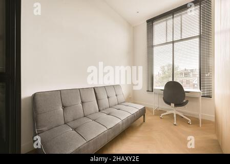 Inneneinrichtung des Zimmers mit weißen Wänden und Laminatboden sowie moderner Arbeitsplatz mit Bürostuhl und Schreibtisch und einem großen Schlafsofa Stockfoto