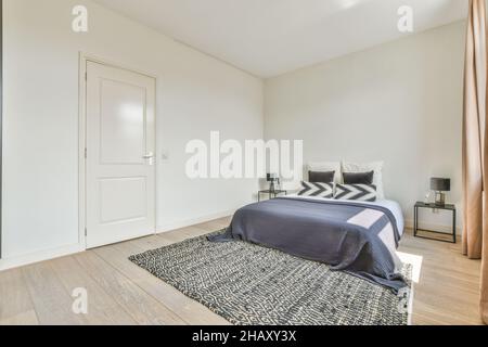 Komfortables Bett mit blauer Decke und Kissen auf Teppich, saubere Fenster mit Vorhängen im geräumigen Schlafzimmer mit Sonnenlicht im Apartment Stockfoto