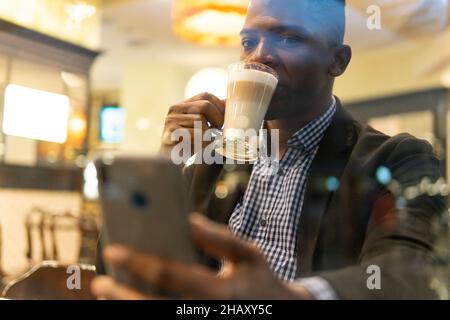 Durch die Fenster eines selbstbewussten jungen afroamerikanischen Unternehmers in elegantem Outfit, der ein Glas Latte Macchiato trinkt und auf dem mobilen Phon Nachrichten übergibt Stockfoto