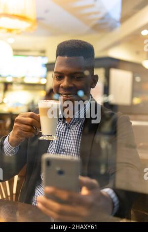 Durch die Fenster eines selbstbewussten jungen afroamerikanischen Unternehmers in elegantem Outfit, der ein Glas Latte Macchiato trinkt und auf dem mobilen Phon Nachrichten übergibt Stockfoto
