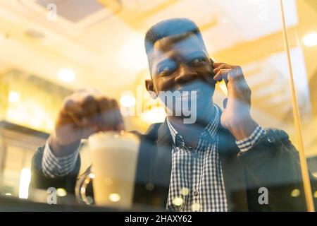 Durch die Fenster des selbstbewussten jungen afroamerikanischen Unternehmers in elegantem Outfit, das Glas Latte Macchiato trinkt und auf einem Telefonat auf Smart Stockfoto