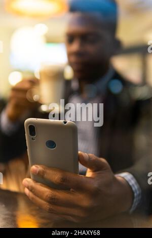 Durch die Fenster eines selbstbewussten jungen afroamerikanischen Unternehmers in elegantem Outfit, der ein Glas Latte Macchiato trinkt und auf dem mobilen Phon Nachrichten übergibt Stockfoto