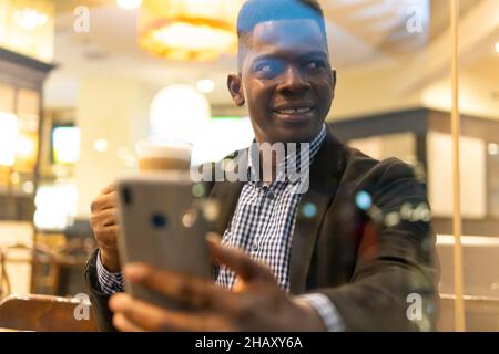 Durch die Fenster eines selbstbewussten jungen afroamerikanischen Unternehmers in elegantem Outfit, der ein Glas Latte Macchiato trinkt und auf dem mobilen Phon Nachrichten übergibt Stockfoto