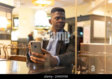 Durch die Fenster eines selbstbewussten jungen afroamerikanischen Unternehmers in elegantem Outfit, der ein Glas Latte Macchiato trinkt und auf dem mobilen Phon Nachrichten übergibt Stockfoto